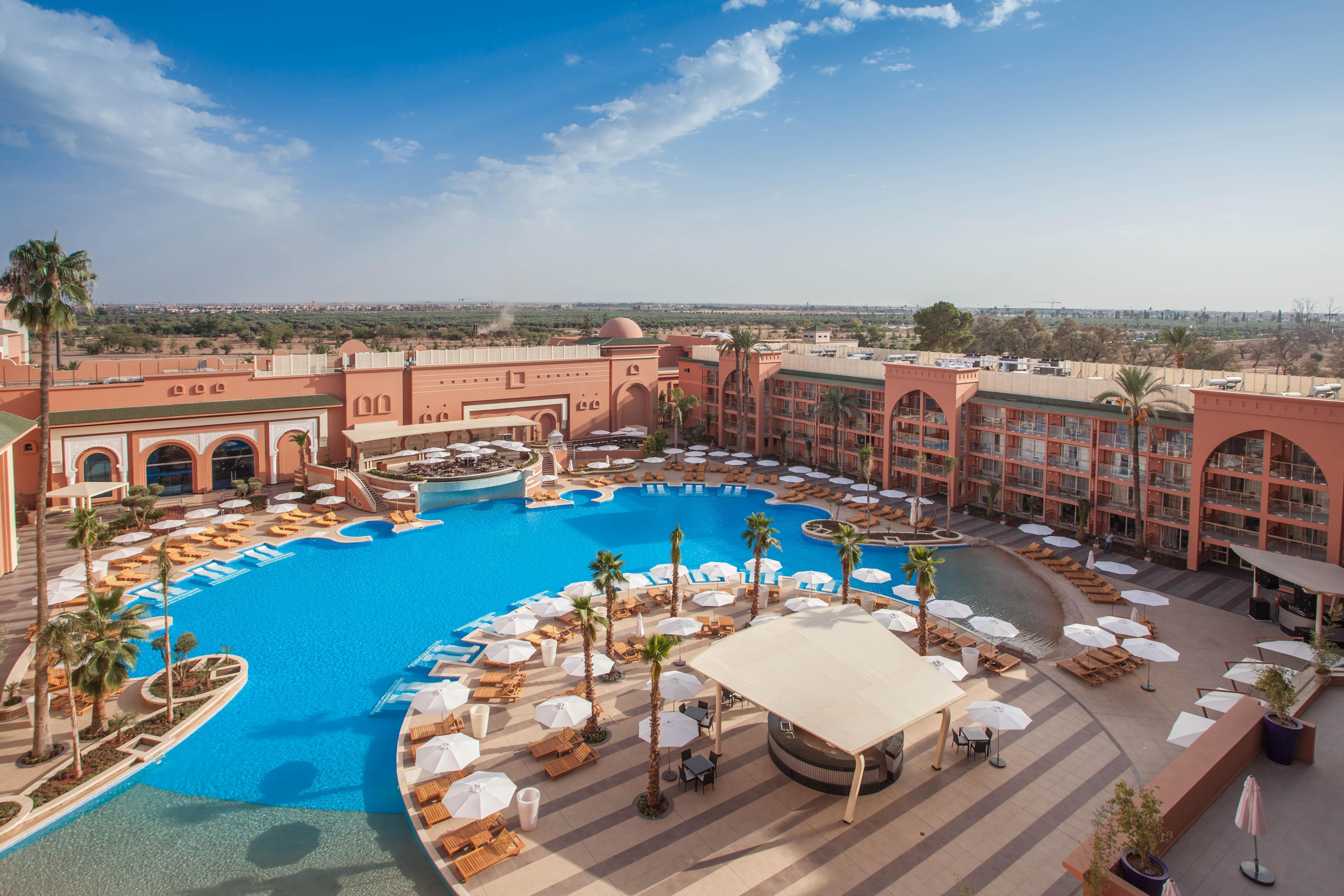 Savoy Le Grand Hotel Marrakech Marrakesz Zewnętrze zdjęcie The photo depicts a luxurious resort featuring a large, inviting swimming pool surrounded by sun loungers and umbrellas. The pool area is designed with a circular layout, and there are palm trees adding a tropical feel. In the background, there are s