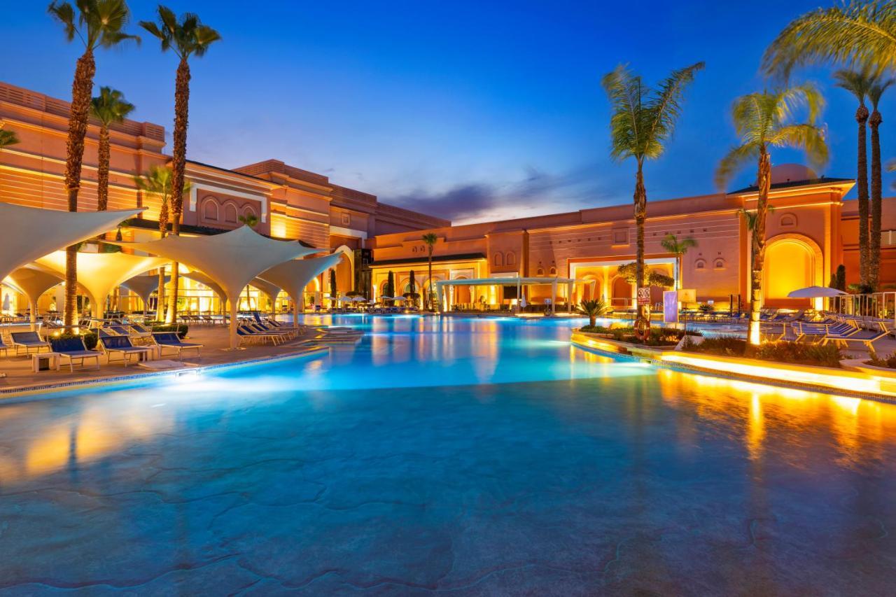 Savoy Le Grand Hotel Marrakech Marrakesz Zewnętrze zdjęcie The photo shows a luxurious resort pool area during twilight. The scene features a large, inviting swimming pool with illuminated edges, reflecting the soft light of the sky. Surrounding the pool are stylish lounge chairs and shade structures. Tall p