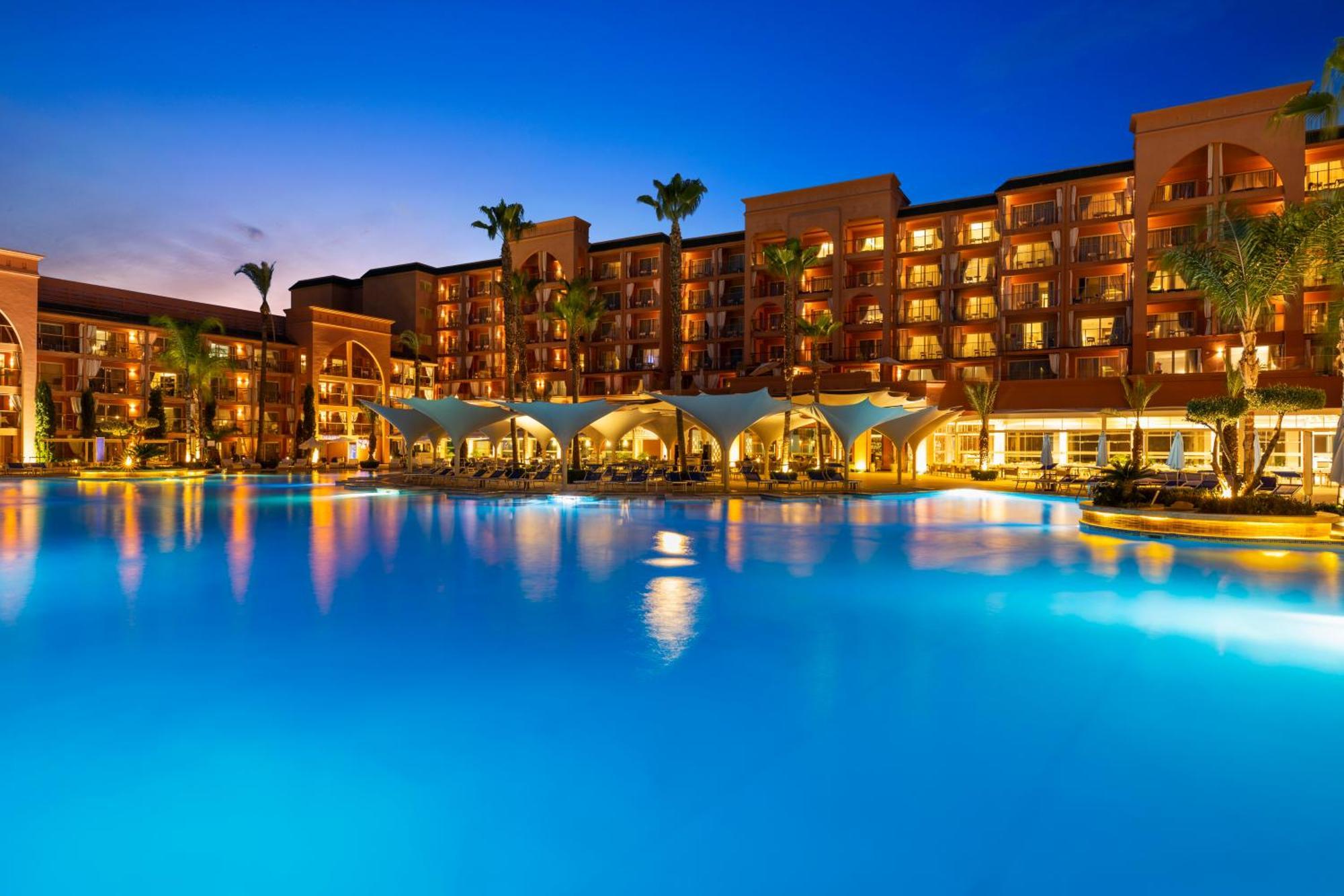 Savoy Le Grand Hotel Marrakech Marrakesz Zewnętrze zdjęcie The photo depicts a luxurious resort setting during twilight. There is a large, illuminated swimming pool in the foreground with a vibrant blue hue, reflecting the evening sky. Surrounding the pool are several palm trees, adding a tropical feel to th