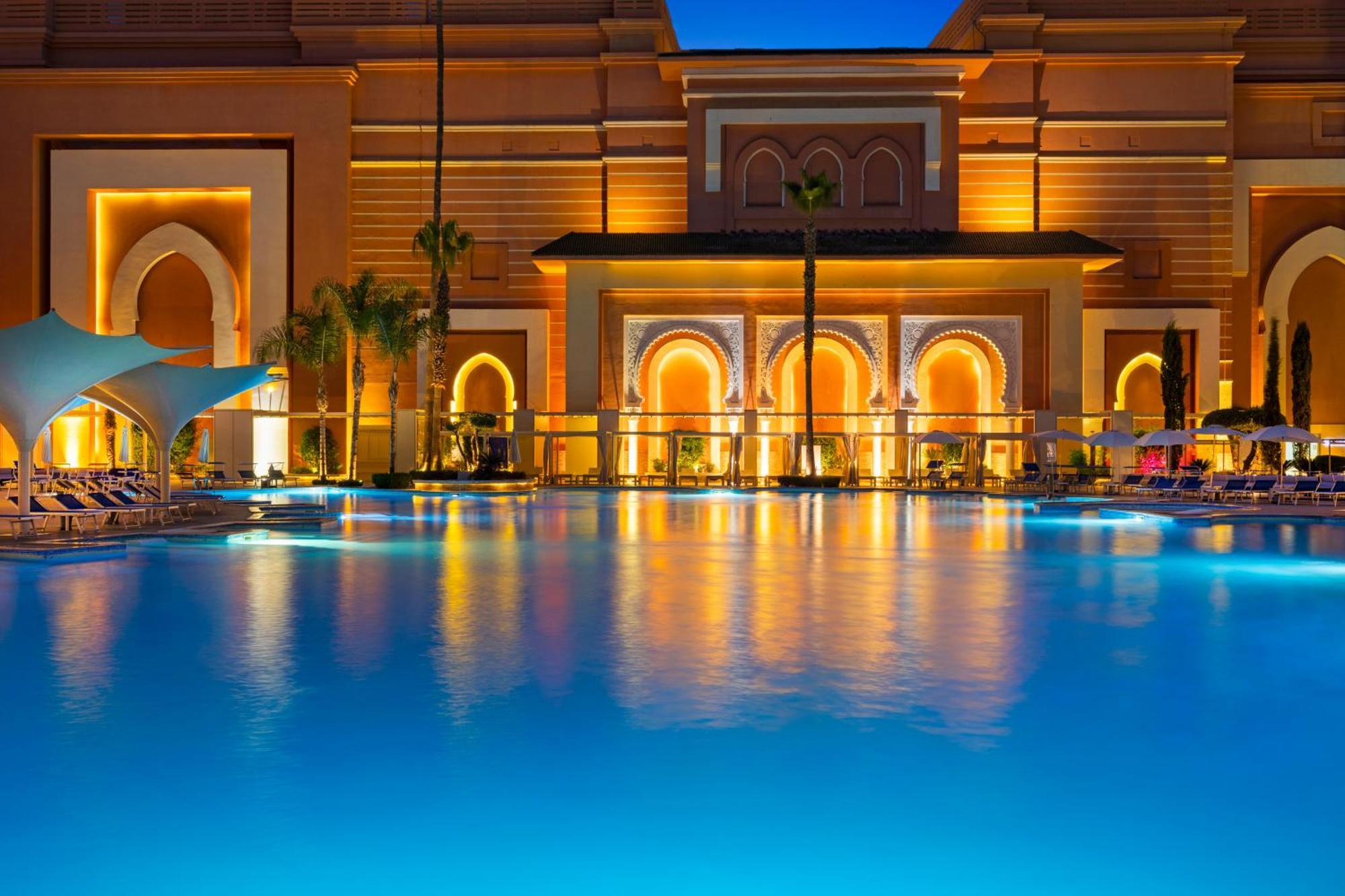 Savoy Le Grand Hotel Marrakech Marrakesz Zewnętrze zdjęcie The photo shows a beautifully illuminated resort at dusk. In the foreground, a swimming pool reflects the blue of the water and the warm lighting of the surrounding architecture. The buildings in the background feature intricate designs with arched d