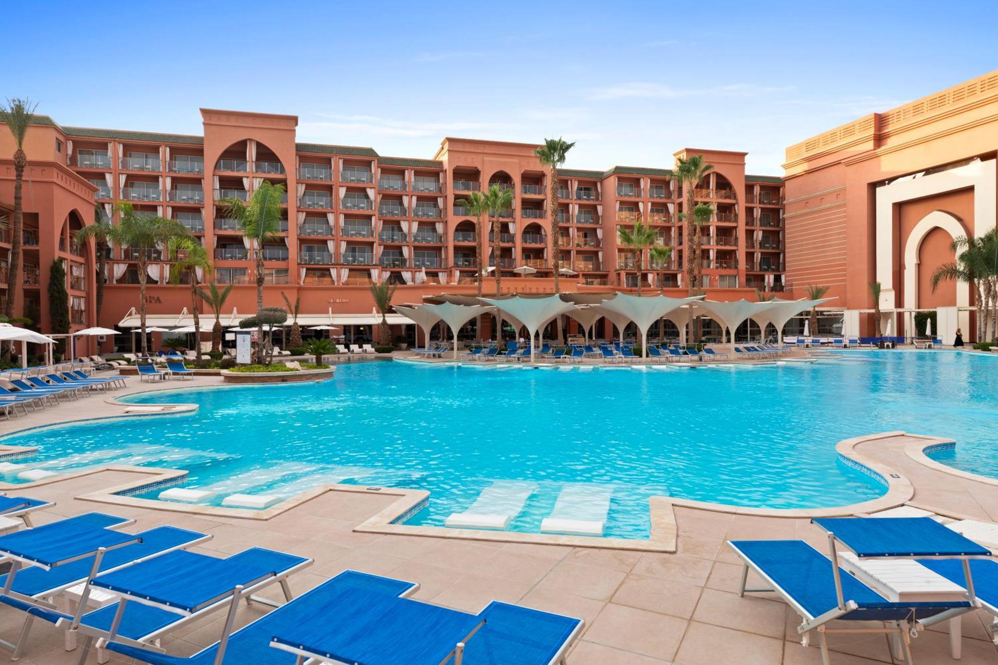 Savoy Le Grand Hotel Marrakech Marrakesz Zewnętrze zdjęcie The image shows a luxurious hotel setting featuring a large swimming pool in the foreground. The pool is surrounded by lounge chairs, primarily in blue, and has several shaded areas underneath canopies. In the background, there is a multi-story hotel