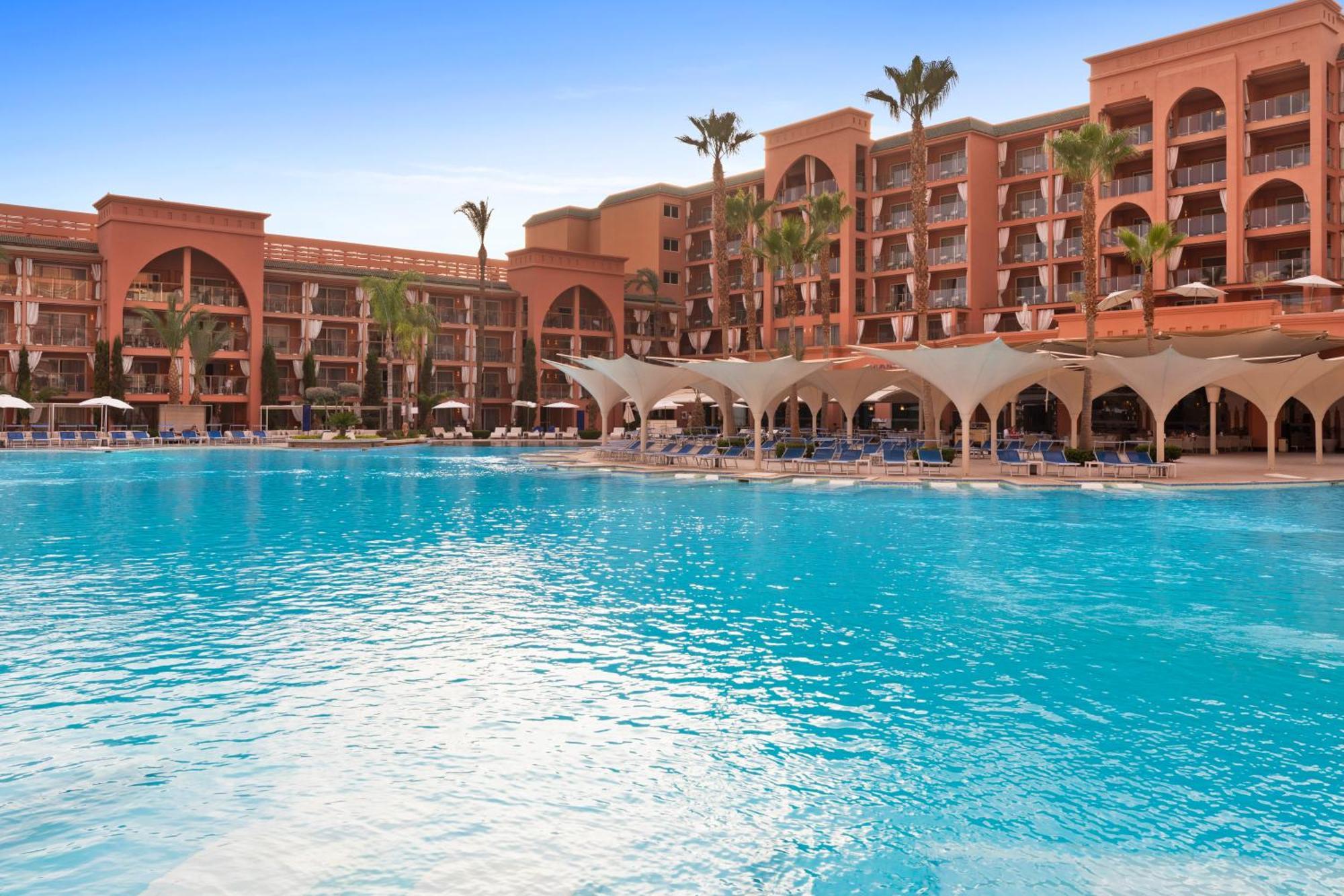 Savoy Le Grand Hotel Marrakech Marrakesz Zewnętrze zdjęcie The photo shows a luxurious hotel or resort with a large, inviting swimming pool in the foreground. The pool features a vibrant blue color and is surrounded by comfortable lounge chairs and shaded areas. In the background, there are multiple building