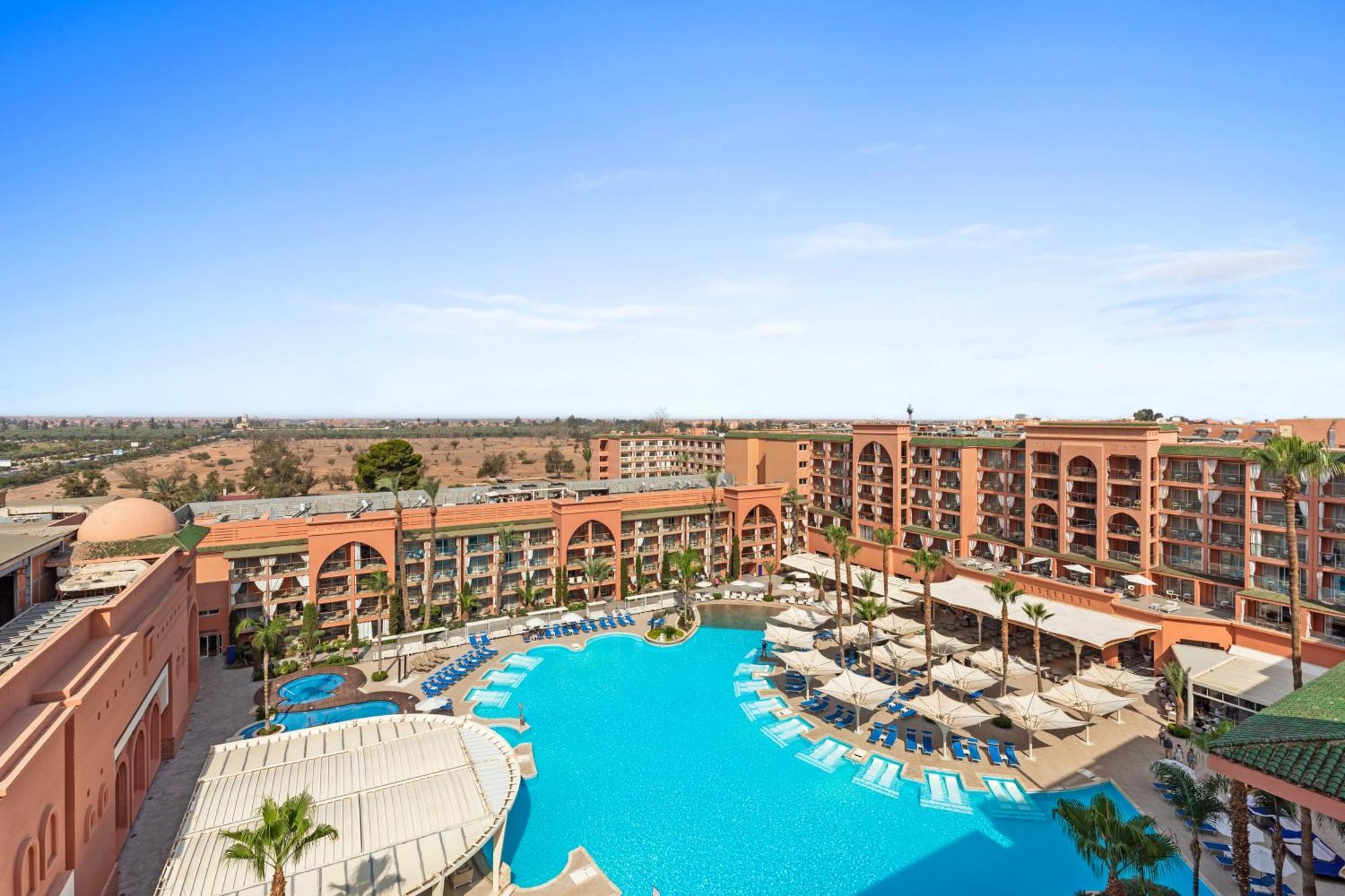 Savoy Le Grand Hotel Marrakech Marrakesz Zewnętrze zdjęcie The image shows a resort with a large swimming pool in the foreground, surrounded by lounge chairs and umbrellas. The pool area is adorned with palm trees, contributing to a tropical vibe. In the background, there is a multi-story building featuring 