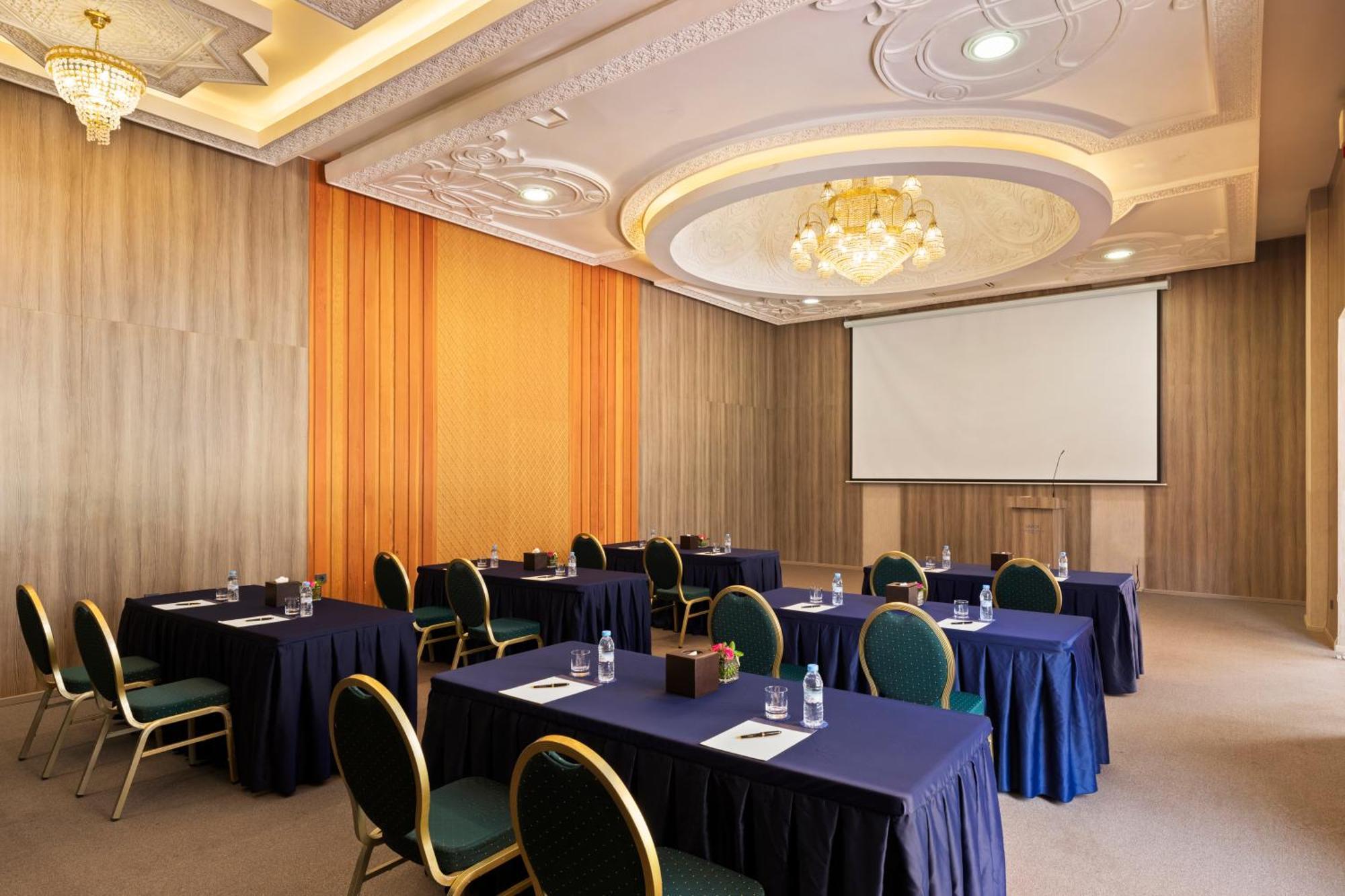 Savoy Le Grand Hotel Marrakech Marrakesz Zewnętrze zdjęcie The photo shows a conference or meeting room. It features several tables arranged in a classroom or seminar style, each covered with a dark blue tablecloth. There are green chairs positioned around the tables. On the tables, there are water bottles a