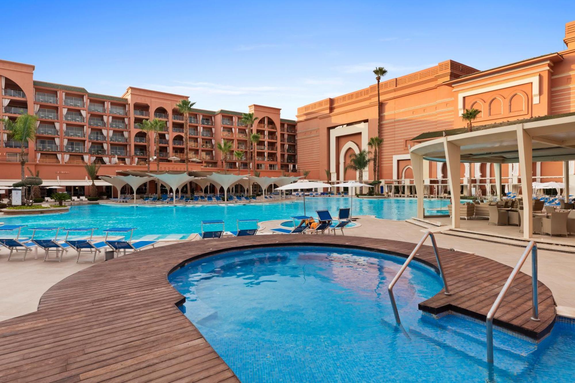 Savoy Le Grand Hotel Marrakech Marrakesz Zewnętrze zdjęcie The photo showcases a vibrant resort with a large swimming pool and a smaller jacuzzi area. The pool area is surrounded by lounge chairs, providing space for relaxation. In the background, there are several multi-story buildings designed with a Moroc