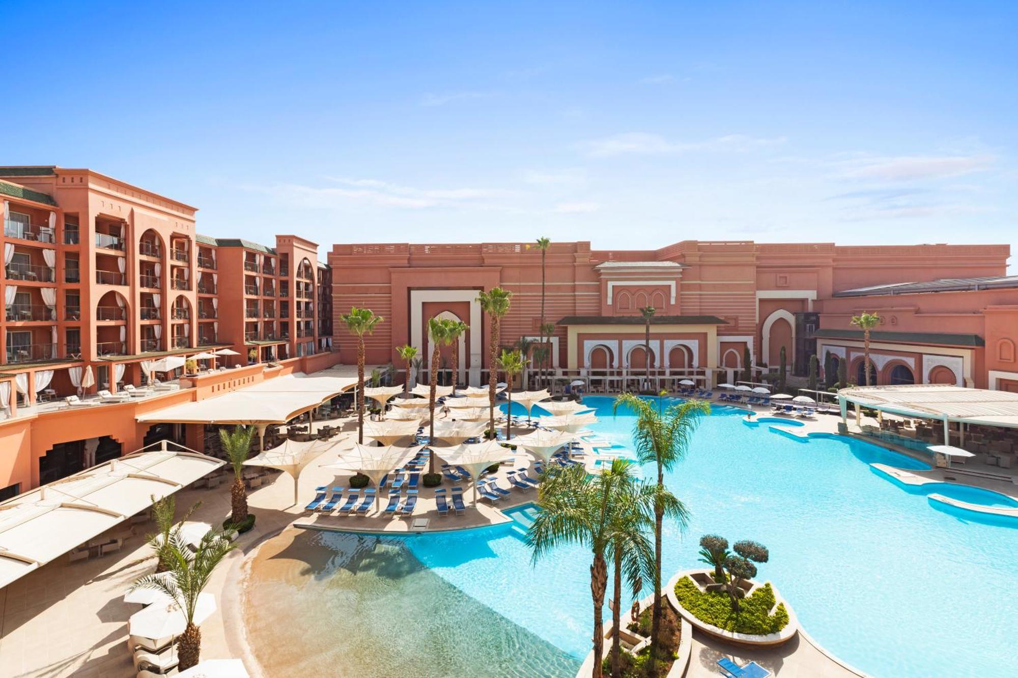 Savoy Le Grand Hotel Marrakech Marrakesz Zewnętrze zdjęcie The photo shows a large, luxurious hotel or resort with a prominent swimming pool in the foreground. The pool area features lounge chairs and palm trees, creating a tropical atmosphere. The hotel itself has an elegant architectural design, characteri