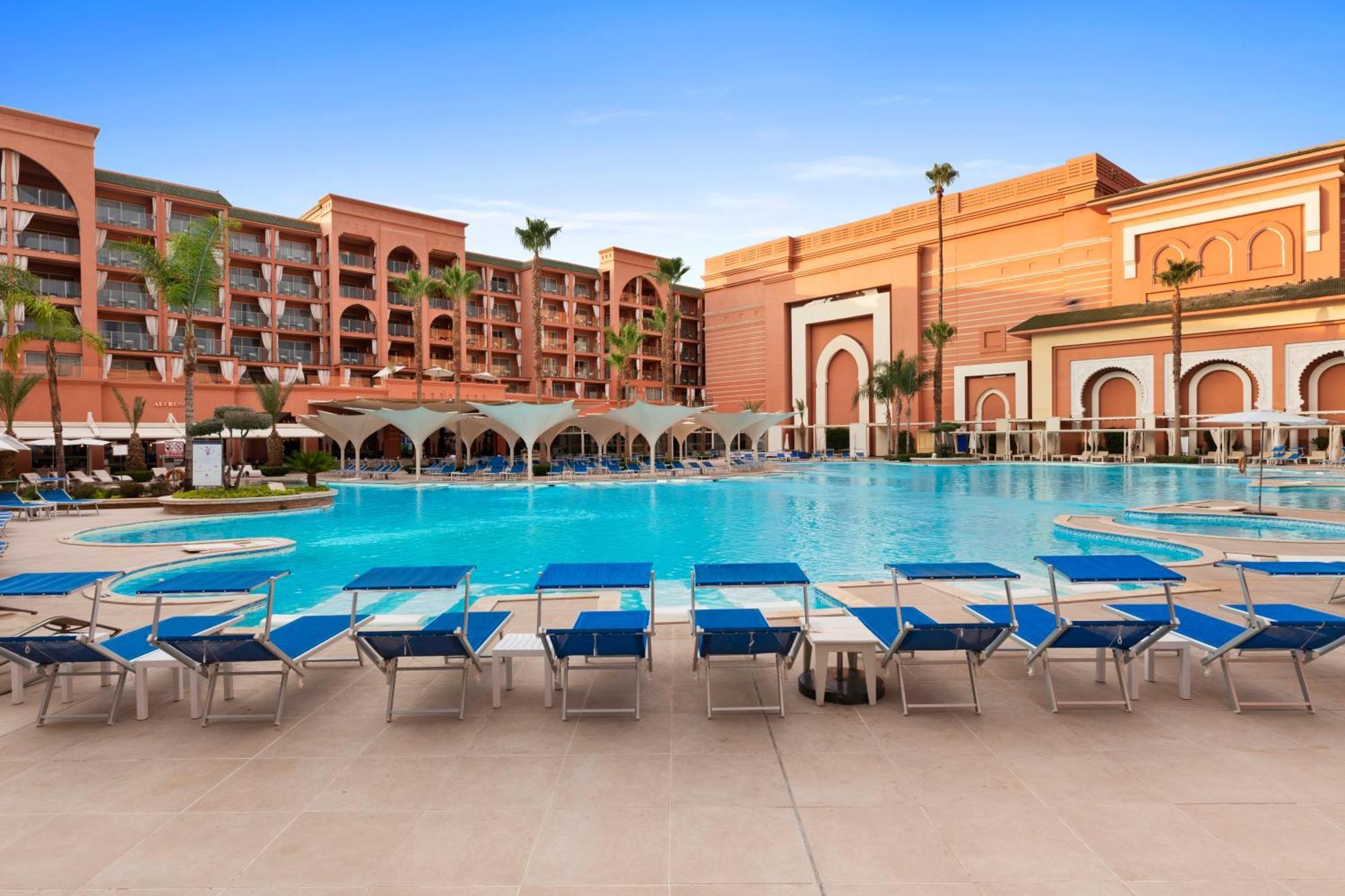 Savoy Le Grand Hotel Marrakech Marrakesz Zewnętrze zdjęcie The image shows a hotel or resort setting featuring a large swimming pool. The pool area is surrounded by lounge chairs that have blue cushions. In the background, there are several buildings with a desert-inspired architectural style, characterized 