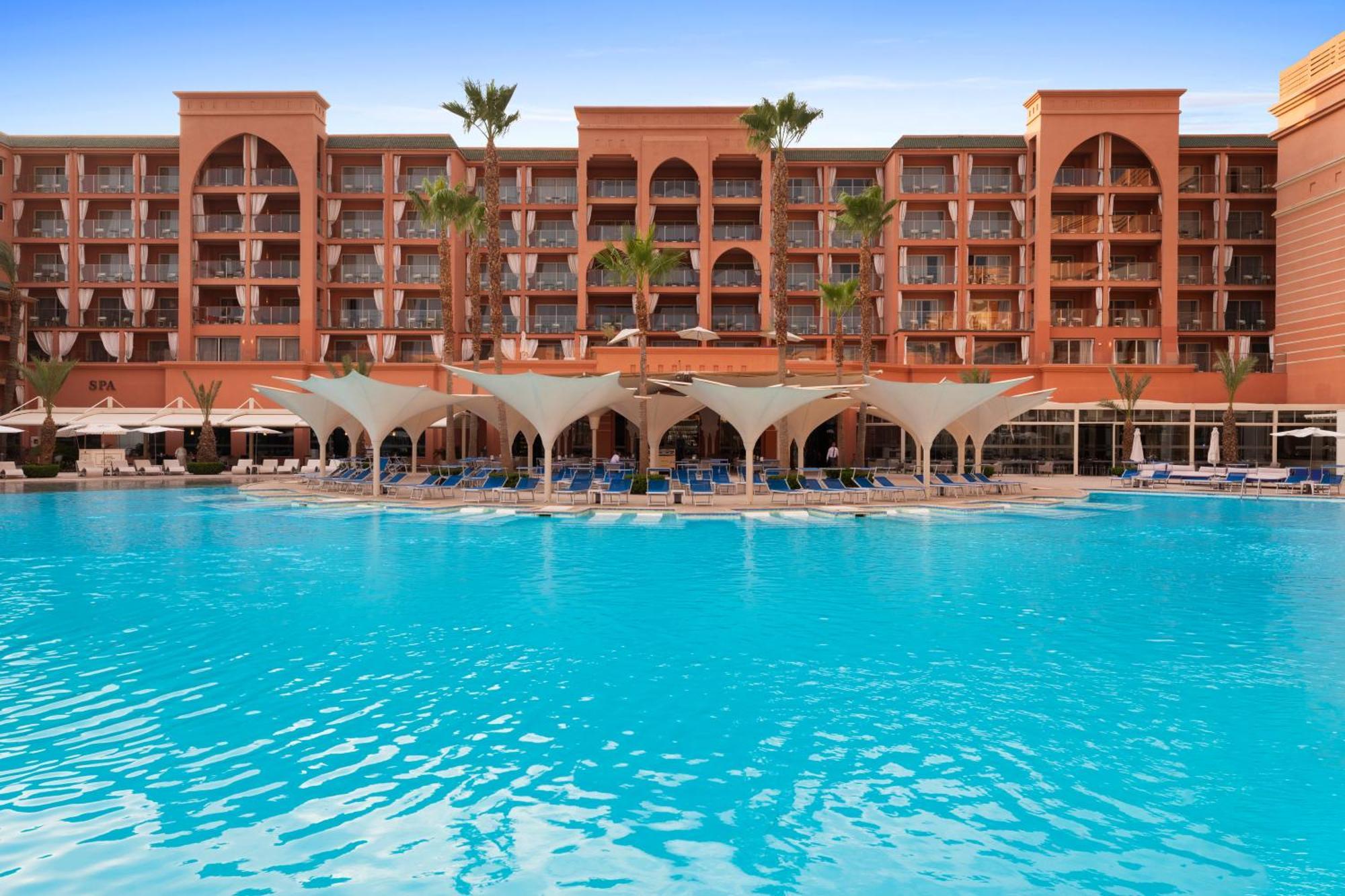 Savoy Le Grand Hotel Marrakech Marrakesz Zewnętrze zdjęcie The photo shows a beautiful hotel with an architectural style typical of Moroccan design, characterized by its red-orange facade and arched windows. In the foreground, there is a large swimming pool with crystal-clear blue water. The pool area is sha