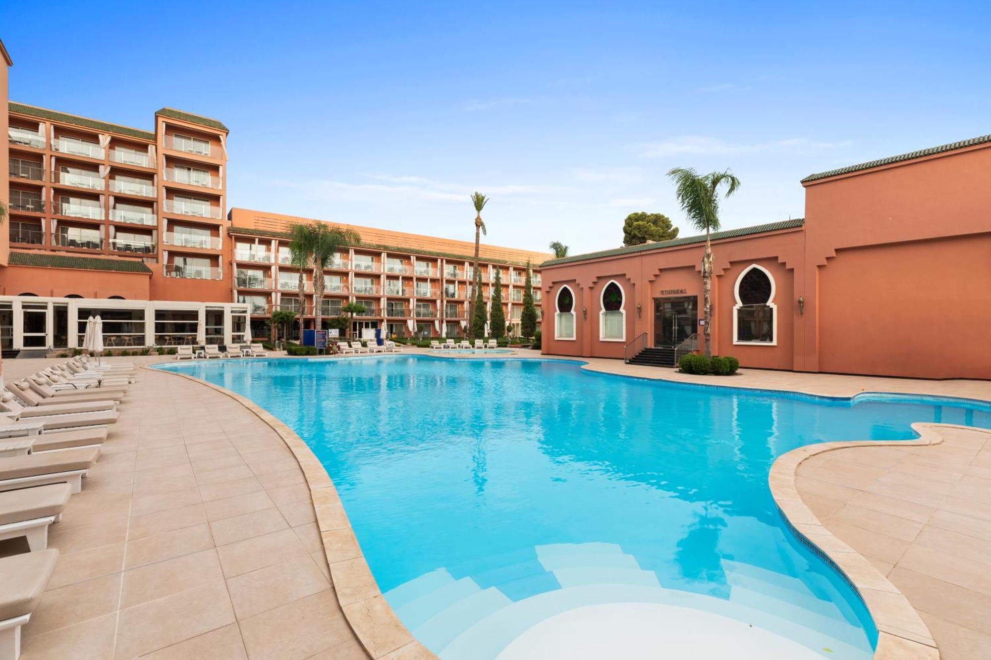 Savoy Le Grand Hotel Marrakech Marrakesz Zewnętrze zdjęcie The photo shows a large swimming pool in an outdoor setting, surrounded by palm trees. On one side of the pool, there is a modern building with multiple floors and balconies, while on the opposite side, there is a structure with a distinctive archite