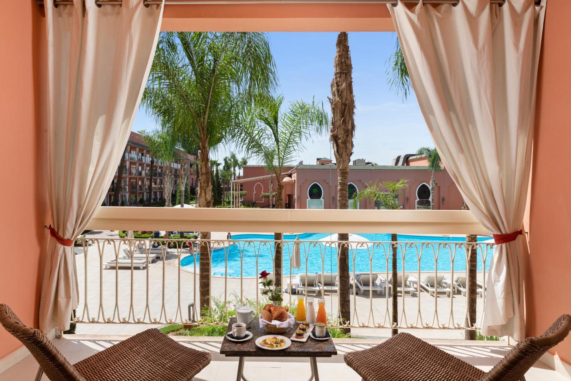 Savoy Le Grand Hotel Marrakech Marrakesz Zewnętrze zdjęcie The photo depicts a view from a balcony overlooking a swimming pool area. There are palm trees surrounding the pool, and several lounge chairs are arranged near the water. In the foreground, two wicker chairs are set up at a small table, which is ado