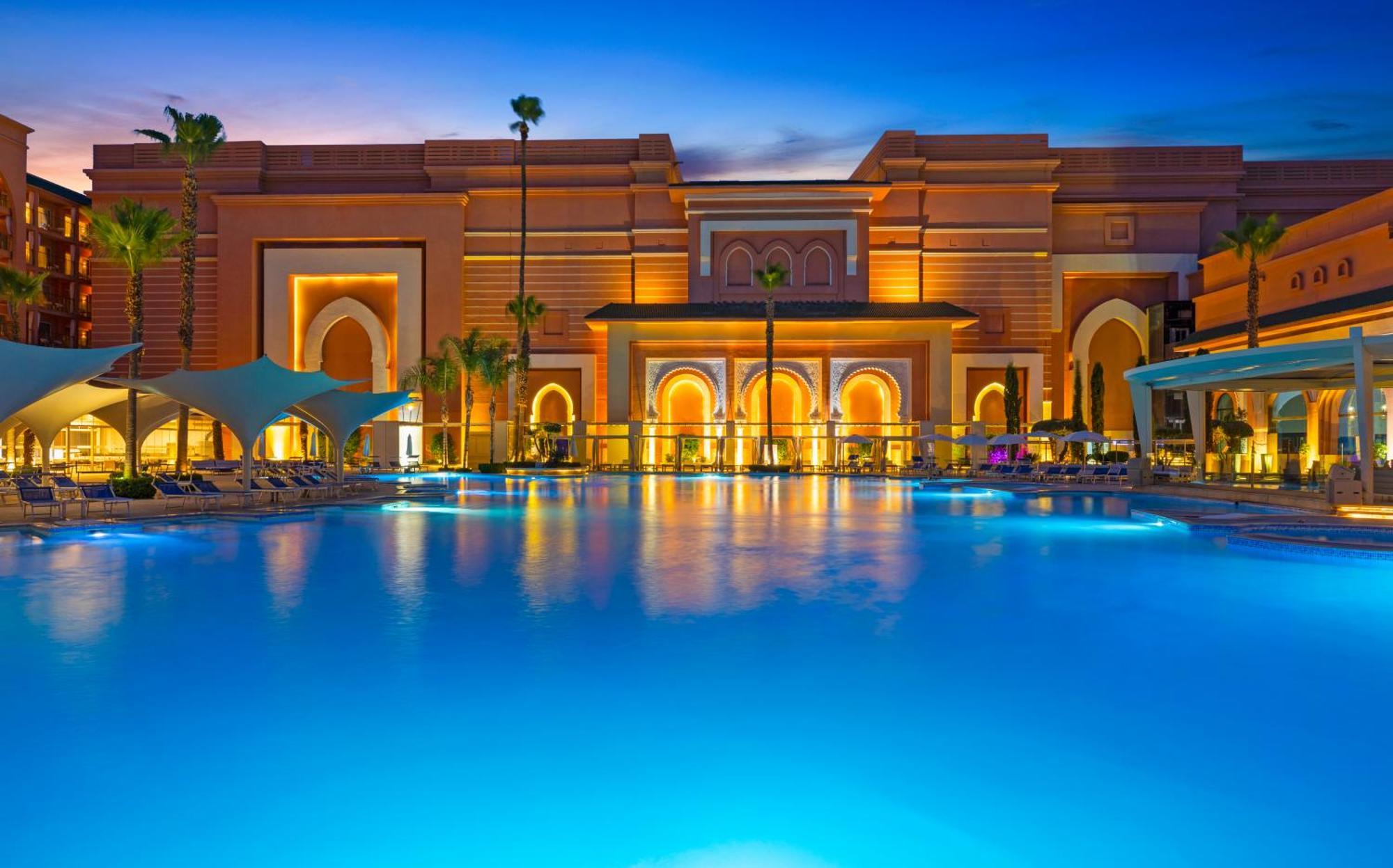 Savoy Le Grand Hotel Marrakech Marrakesz Zewnętrze zdjęcie The photo depicts a luxurious resort featuring a large, illuminated swimming pool in the foreground. The pool is surrounded by palm trees and lounge chairs, reflecting vibrant blue water. In the background, there is an elaborate building designed wit