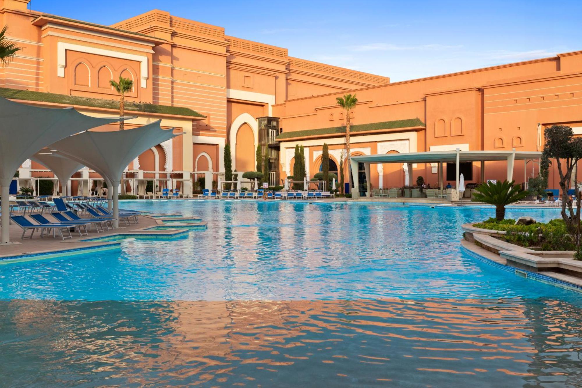 Savoy Le Grand Hotel Marrakech Marrakesz Zewnętrze zdjęcie The photo shows a large, inviting swimming pool with clear blue water, surrounded by lounge chairs. In the background, there is an elegantly designed building with a warm-colored facade, featuring arches and decorative elements, typical of luxury res